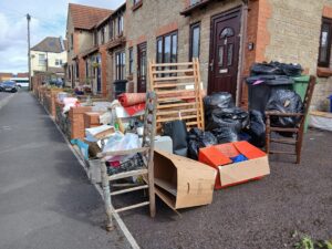 A pile of illegally dumped fly-tip on a pavement. There are bags, boxes and broken furniture horrible in the  pile.