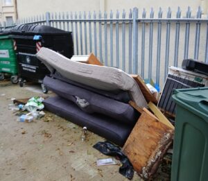 A fly-tip by a fence. Old, manky mattresses and other household items are left in a pile next to some bins