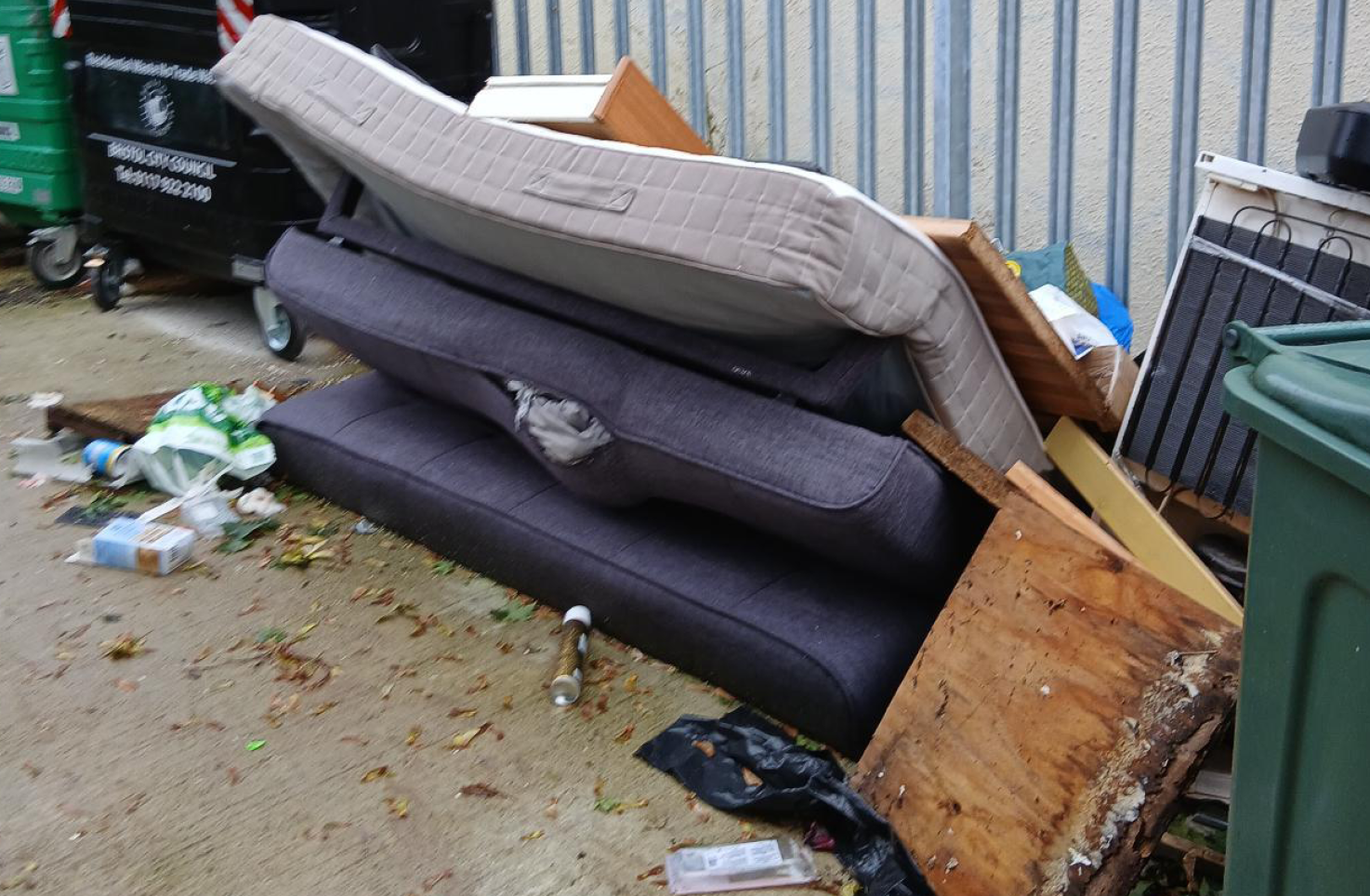 Old, manky mattresses and other household items are left in a pile next to some bins