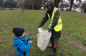 A child uses a litter picking stick to put something into a bin bag, held open by a grown up in a hi-vis vest